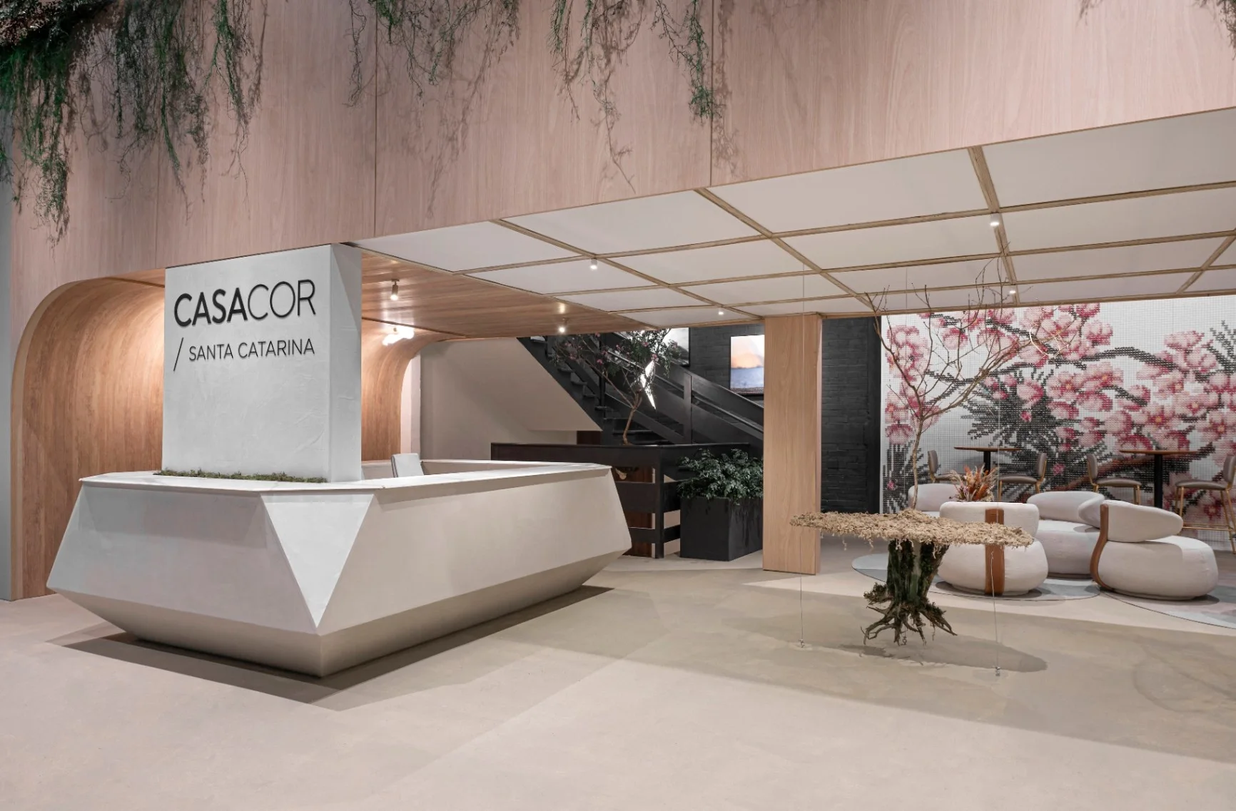 Recepção do ambiente Ikigai na CASACOR Santa Catarina, com balcão facetado, arco em madeira e painel de flores de cerejeira ao fundo